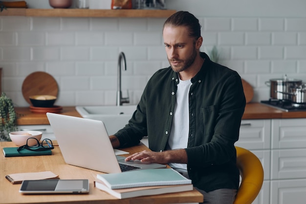 Zekerzinnige jonge man die een laptop gebruikt terwijl hij thuis aan de keukentafel werkt