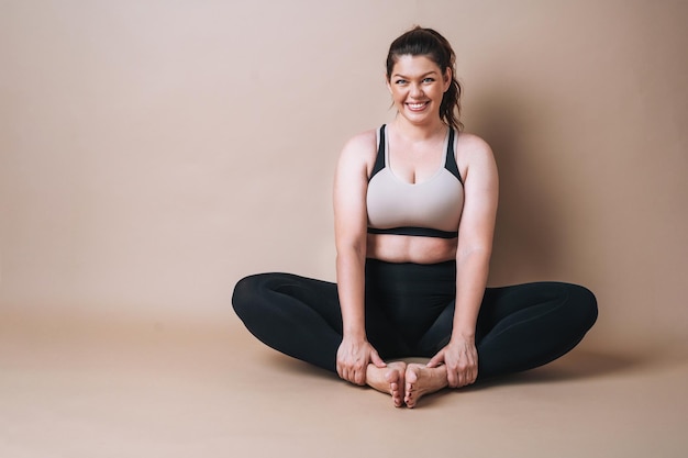 Zelf liefhebbende jonge vrouw plus maat in sportkleding die zich uitstrekt op beige achtergrond lichaamsliefde