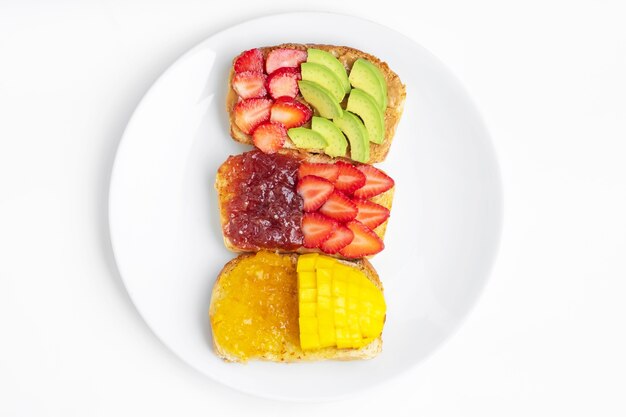 Zelfgebakken brood bedekt met pindakaas, sinaasappeljam en aardbeienjamtop met aardbeien, mango en avocado op witte plaat. gezonde voeding voor gewichtsverlies. gezond ontbijtconcept.