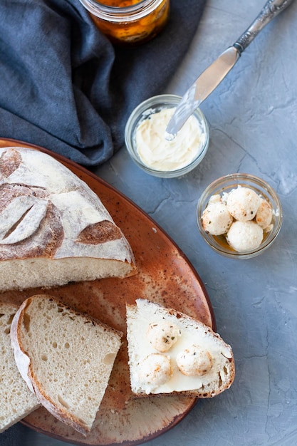 Zelfgebakken brood half en plakjes met botervloot en mes, glazen pot en gerookte kaas op grijze achtergrond