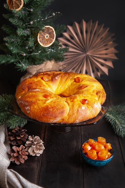 Zelfgebakken kerstbrood in een vorm van adventskrans van gistdeeg met oranje gekonfijte vruchten op de achtergrond van de kerstboom.