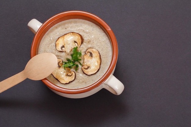 Zelfgemaakte champignonroomsoep in porseleinen kom met gesneden champignons en houten lepel op een zwarte stenen bord. Bovenaanzicht.