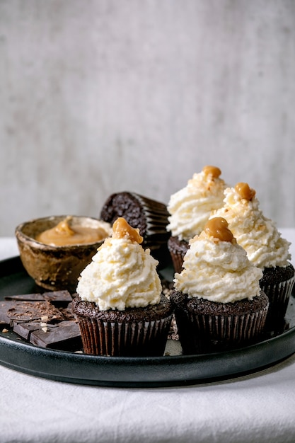 Zelfgemaakte chocolade cupcakes muffins met witte slagroom botercrème en gezouten karamel, geserveerd met gehakte donkere chocolade op zwarte keramische plaat op wit tafelkleed.