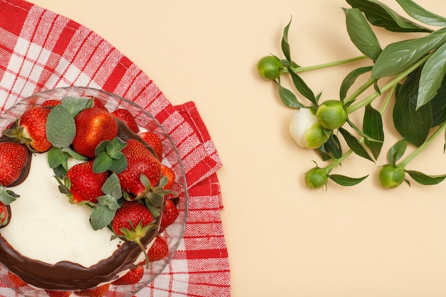 Zelfgemaakte chocoladetaart versierd met verse aardbeien en blaadjes munt op glasplaat met keukenservet en boeket pioenrozen op beige kleur achtergrond. Bovenaanzicht