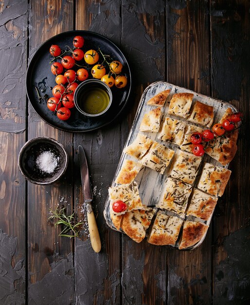 Zelfgemaakte focaccia plat brood in stukjes gesneden geserveerd met gebakken tomaten, olijfolie en verse rozemarijn, met vintage servet en mes op een houten achtergrond. Bovenaanzicht