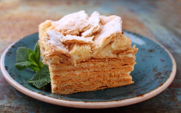 Foto zelfgemaakte gelaagde napoleon-cake met melkroom versierd met munt, op een bord op een rustieke achtergrond.