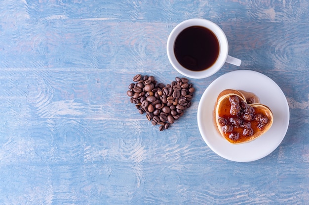 Zelfgemaakte hartvormige pannenkoeken met bessenjam, hartvorm gemaakt van koffiebonen en een witte kop warme koffie op een blauwe houten tafel