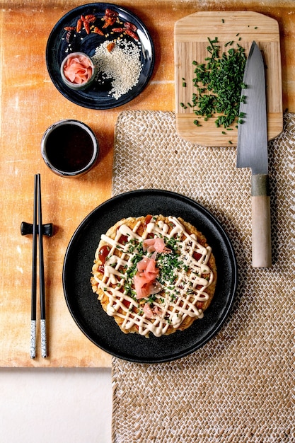 Zelfgemaakte Japanse fastfood okonomiyaki kool pannenkoek versierd met lente-ui, ingelegde gember, mayo saus op zwarte keramische plaat met stokjes en ingrediënten hierboven. Textuur tafel. Plat leggen