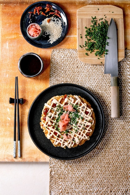 Zelfgemaakte japanse fastfood okonomiyaki kool pannenkoek versierd met lente-ui, ingelegde gember, mayo saus op zwarte keramische plaat met stokjes en ingrediënten hierboven. textuur tafel. plat leggen