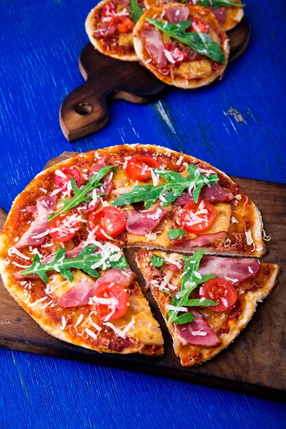 Zelfgemaakte pizza met prosciutto, tomaat, rucola op houten plank blauw oppervlak,