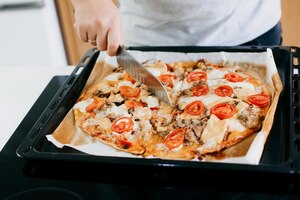 Zelfgemaakte pizza persoon die gekookte verse pizza snijdt met kaas, tomaat, mozzarella, kip en champignons op moderne witte keuken proces om pizza thuis te koken