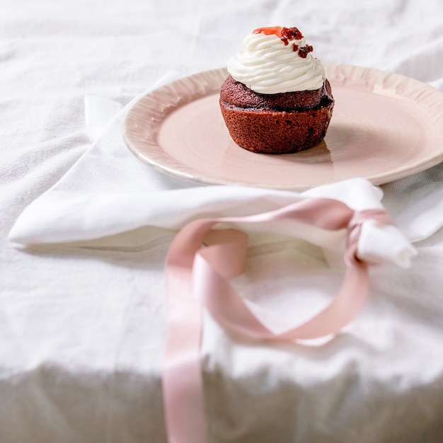 Zelfgemaakte rode fluwelen cupcake met slagroom op roze keramische plaat, wit servet met lint op wit linnen tafelkleed. Ruimte kopiëren. vierkante afbeelding