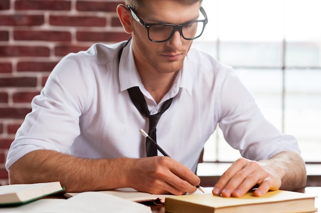 Zelfverzekerd en creatief. Knappe jonge man in overhemd en stropdas die iets in notitieblok schrijft terwijl hij op zijn werkplek zit