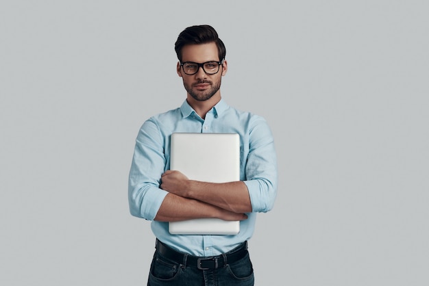 Zelfverzekerd en slim. Knappe jonge man die laptop draagt en glimlacht terwijl hij tegen een grijze achtergrond staat