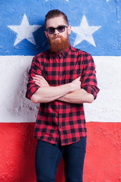 Foto zelfverzekerd en ziet er cool uit. knappe jonge, bebaarde man met een zonnebril die zijn armen gekruist houdt en naar de camera kijkt terwijl hij tegen de amerikaanse vlag staat