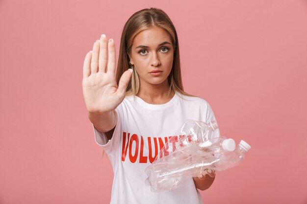 Zelfverzekerd jong vrijwilligersmeisje dat geïsoleerd staat en een stopgebaar toont terwijl ze plastic flessen vasthoudt
