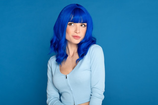 Foto zelfverzekerd jong vrouwelijk model in blauwe, heldere pruik en vrijetijdskleding die dromerig wegkijkt tegen de blauwe achtergrond in de studio