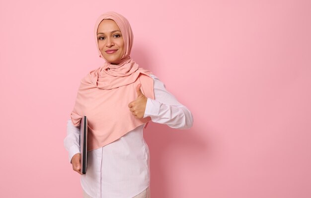 Zelfverzekerd portret van een succesvolle prachtige Arabische moslimvrouw in roze hijab en strikte casual kleding met een duim omhoog, kijkend naar de camera, poserend tegen een gekleurde pastelachtergrond met ruimte voor tekst