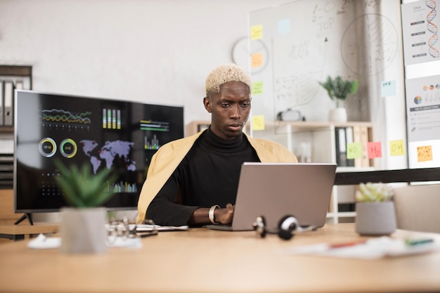 Zelfverzekerde Afrikaanse man in geel pak zittend op kantoor en met behulp van draadloze laptop