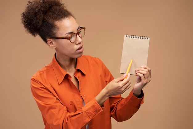 Zelfverzekerde Afrikaanse vrouw in oranje casual shirt schrijft plannen op kladblok geïsoleerde beige achtergrond