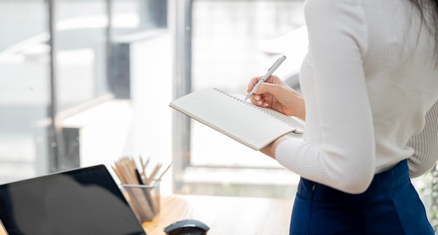 Zelfverzekerde aziatische zakenvrouw die staat en aantekeningen maakt in het financiële boek inkomstenbelasting en werkt op laptop op kantoor