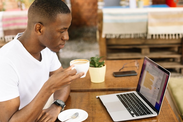 Zelfverzekerde en glimlachende multiraciale zakenman die koffie drinkt die online werkt met behulp van een laptop in een café op afstand