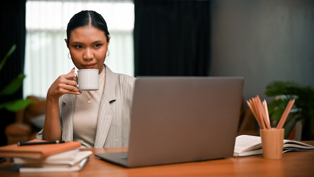 Zelfverzekerde en vastberaden Aziatische zakenvrouw die koffie drinkt terwijl ze op een laptop werkt