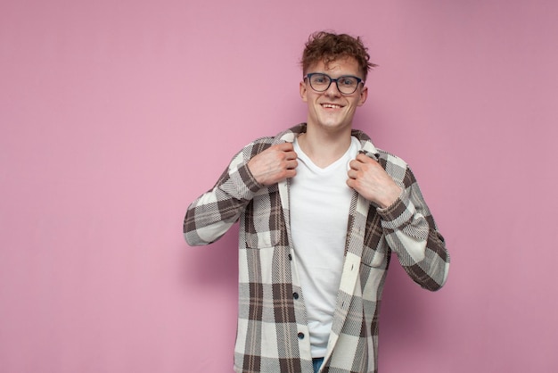 Zelfverzekerde gekrulde man met bril trekt zijn geruite overhemd recht op een roze achtergrond en glimlacht dat een man een overhemd aantrekt