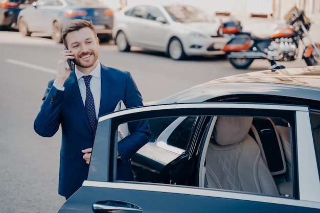 Zelfverzekerde glimlachende directeur gekleed in blauw formeel pak die zwarte limousine verlaat na aankomst op zijn werkplek, telefoneert, krant vasthoudt terwijl hij alleen staat naast auto met open deur