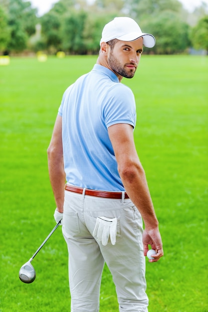 Foto zelfverzekerde golfer. achteraanzicht van zelfverzekerde golfer die golfbal en bestuurder vasthoudt terwijl hij op de golfbaan staat en over de schouder kijkt