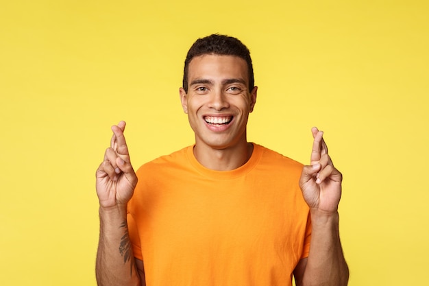 Zelfverzekerde, hoopvolle, vastberaden jonge man in oranje t-shirt, kruisende vingers