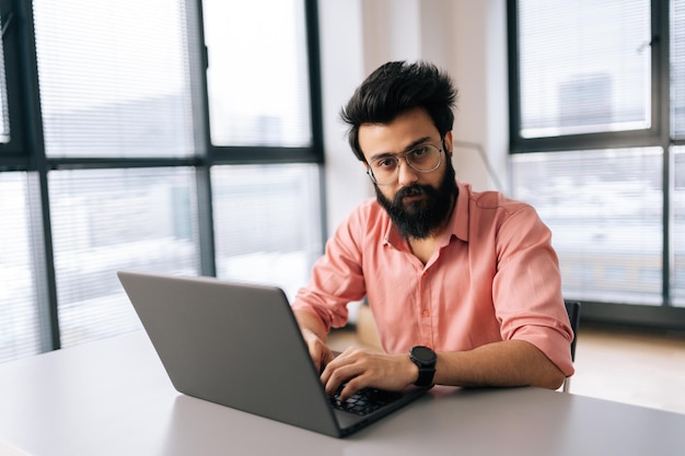 Zelfverzekerde Indiase zakenman in vrijetijdskleding zittend aan een bureau met laptop kijkend naar camera met ernstige uitdrukking in licht coworking-kantoor bij raam Bebaarde freelancer man aan het werk op notebook