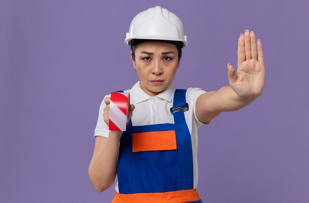 Zelfverzekerde jonge aziatische bouwvrouw met een witte veiligheidshelm die waarschuwingstape vasthoudt en een stopbord gebaren