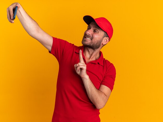 Zelfverzekerde jonge blanke bezorger in rood uniform en pet die vredesteken doet met tanden die selfie nemen geïsoleerd op oranje muur met kopieerruimte