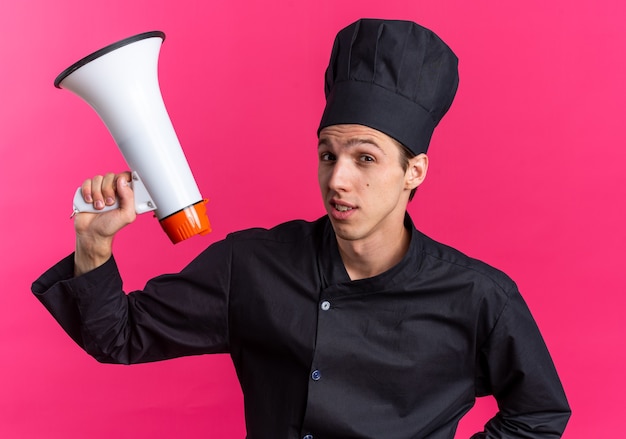 Zelfverzekerde jonge blonde mannelijke kok in uniform van de chef-kok en pet met spreker kijkend naar camera geïsoleerd op roze muur