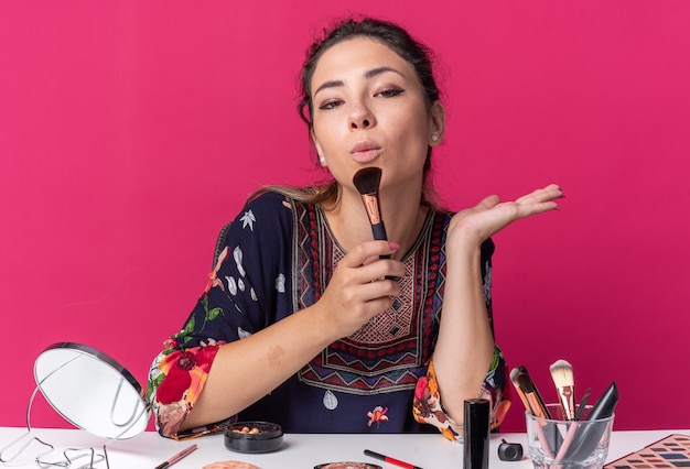 Foto zelfverzekerde jonge brunette meisje zit aan tafel met make-up tools met make-up borstel geïsoleerd op roze muur met kopieerruimte