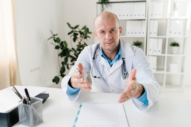 Zelfverzekerde jonge dokter in whitecoat en bril iets aan u uit te leggen zittend op de werkplek