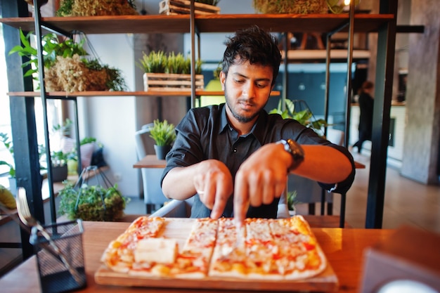 Zelfverzekerde jonge Indiase man in zwart shirt zit bij pizzeria met pizza