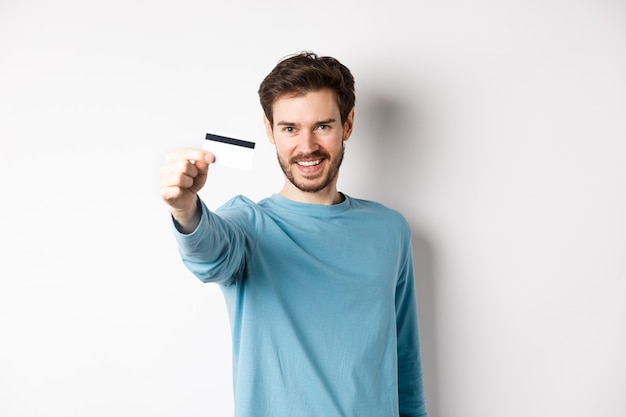 Zelfverzekerde jonge man die lacht, hand uitsteekt en plastic creditcard toont, staande op een witte achtergrond
