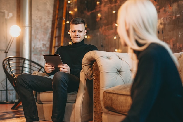 Zelfverzekerde jonge man zittend op een bank en houdt een tablet terwijl het kijken naar haar vriendin lacht die op de vloer zit.