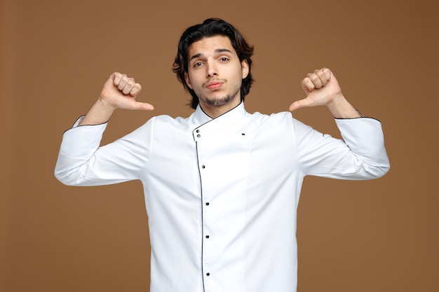 Zelfverzekerde jonge mannelijke chef-kok in uniform kijkend naar camera wijzend op zichzelf geïsoleerd op bruine achtergrond