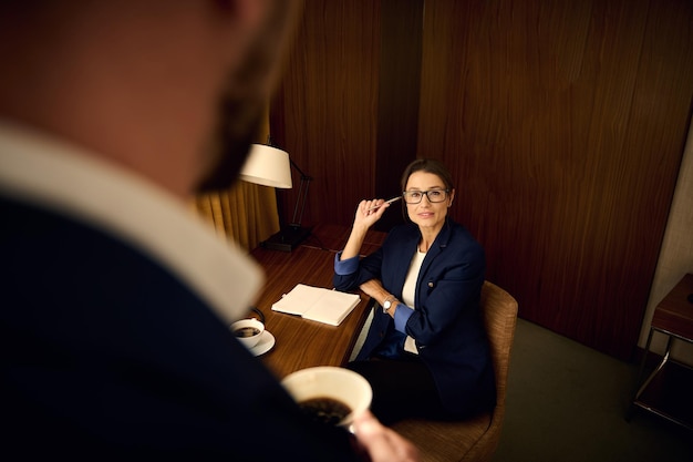 Zelfverzekerde jonge mooie zakenvrouw, vrouwelijke baas, ondernemer zittend aan een tafel en schrijven op notitieblok, vol vertrouwen kijken naar een wazig onherkenbare man in pak op de voorgrond