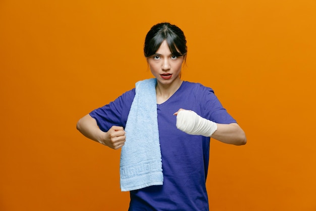 Foto zelfverzekerde jonge sportieve vrouw die t-shirt draagt dat naar camera kijkt die boksgebaar met handdoek op haar schouder toont met pols omwikkeld met verband geïsoleerd op oranje achtergrond