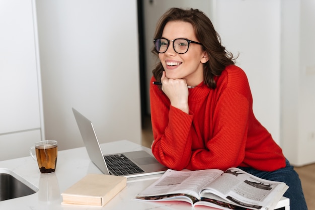 Zelfverzekerde jonge vrouw zittend aan de keukentafel thuis, studeren met laptopcomputer en werkmap