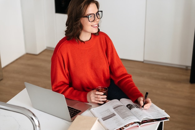 Zelfverzekerde jonge vrouw zittend aan de keukentafel thuis, studeren met laptopcomputer en werkmap