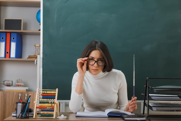 zelfverzekerde jonge vrouwelijke leraar die een bril draagt met de aanwijzer aan het bureau met schoolhulpmiddelen in de klas