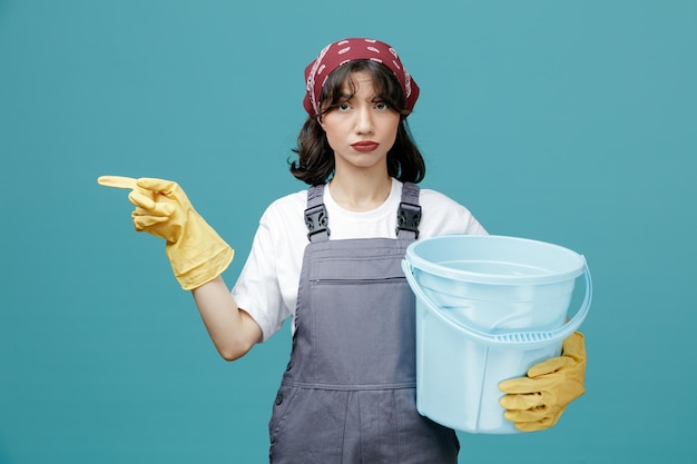 Zelfverzekerde jonge vrouwelijke schoonmaker met uniforme bandana en rubberen handschoenen met emmer kijkend naar camera wijzend naar kant geïsoleerd op blauwe achtergrond