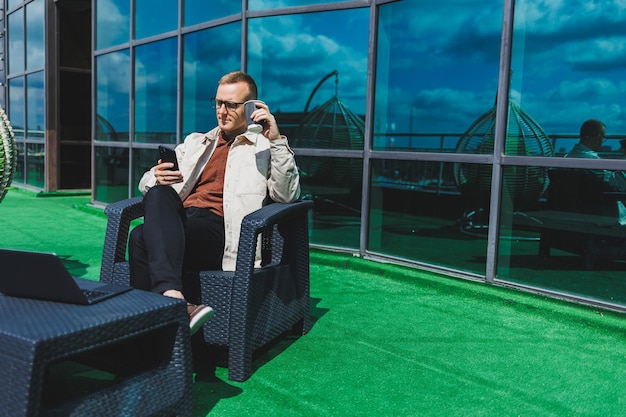 Zelfverzekerde jonge zakenman gekleed in formele kleding en stijlvolle bril roept een smartphone op terwijl hij met koffie zit op het terras van een modern kantoorgebouw met een glazen wand