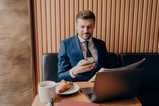 Zelfverzekerde jonge zakenman in formele kleding die van koffie geniet en probeert het drukke werkschema op zijn smartphone bij te houden, op laptop werkt en de krant leest terwijl hij in de coffeeshop zit