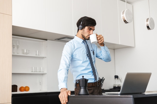 Zelfverzekerde jonge zakenman thuis werken in de keuken, met behulp van laptopcomputer, communiceren via hoofdtelefoon set